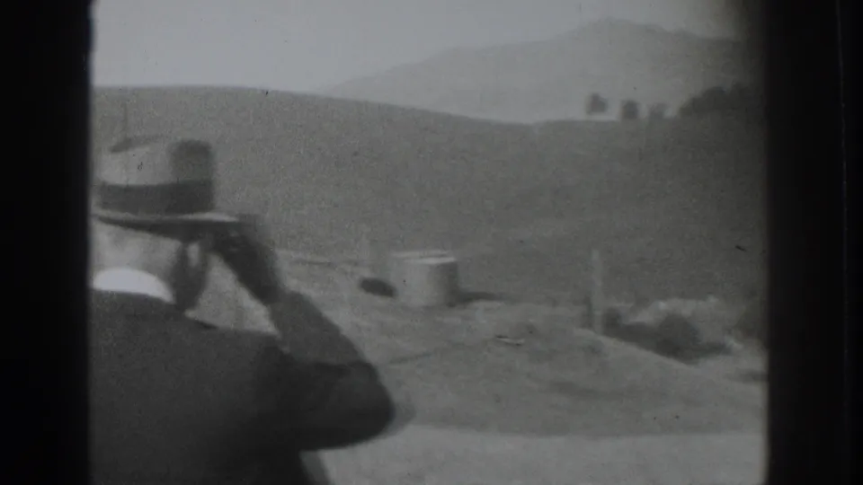 1937 Vacaville home-movie frame showing a suited man in a fedora looking through binoculars across a rural hillside.