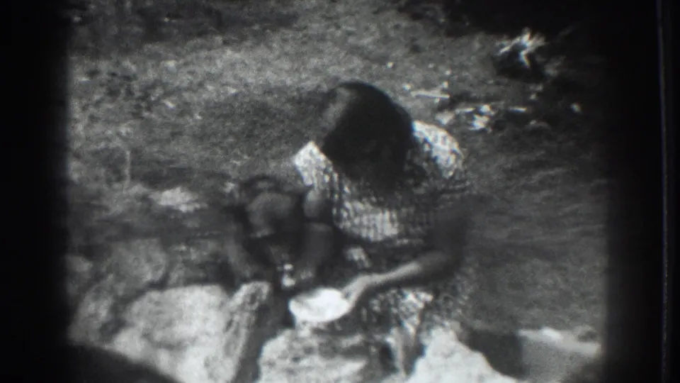 1937 Vacaville home-movie frame of a woman crouching in a rural yard while offering food from a bowl to a dark-furred dog.