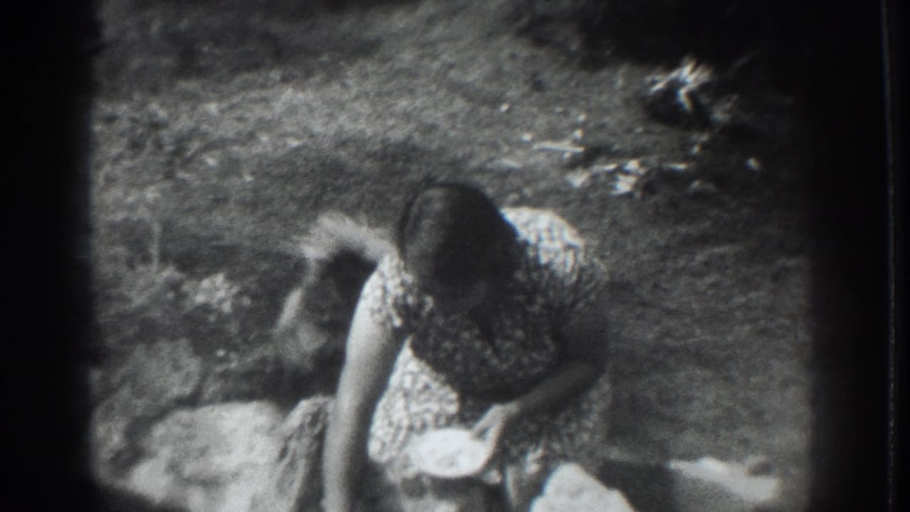 In 1937 Vacaville home-movie footage, a woman in a patterned dress bends toward a small dog beside rocks and garden ground.