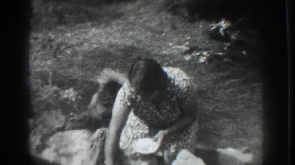 In 1937 Vacaville home-movie footage, a woman in a patterned dress bends toward a small dog beside rocks and garden ground.