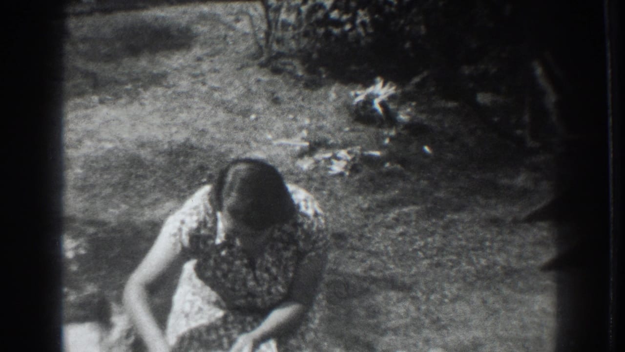 The woman crouches or bends in the garden area while the small dog is partly visible near the right side of the 8mm frame.
