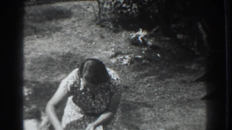 The woman crouches or bends in the garden area while the small dog is partly visible near the right side of the 8mm frame.