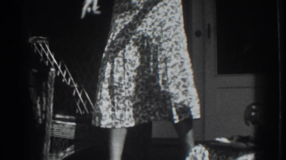 1937 Vacaville home-movie frame showing a woman in a patterned dress beside a wicker chair and house doorway.