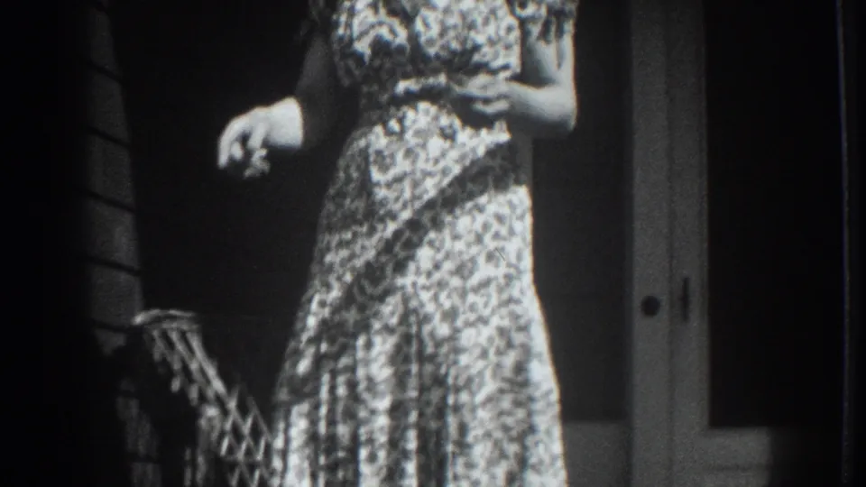 1937 Vacaville frame with a woman in a patterned dress holding a small possible animal or object near a porch doorway.