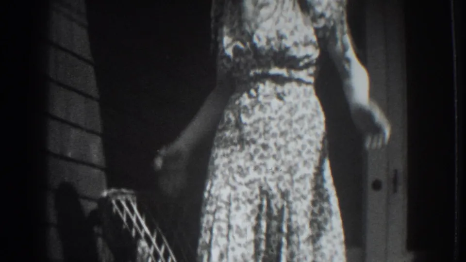 1937 Vacaville porch scene with a woman in a patterned dress standing beside a doorway in grainy monochrome 8mm footage.
