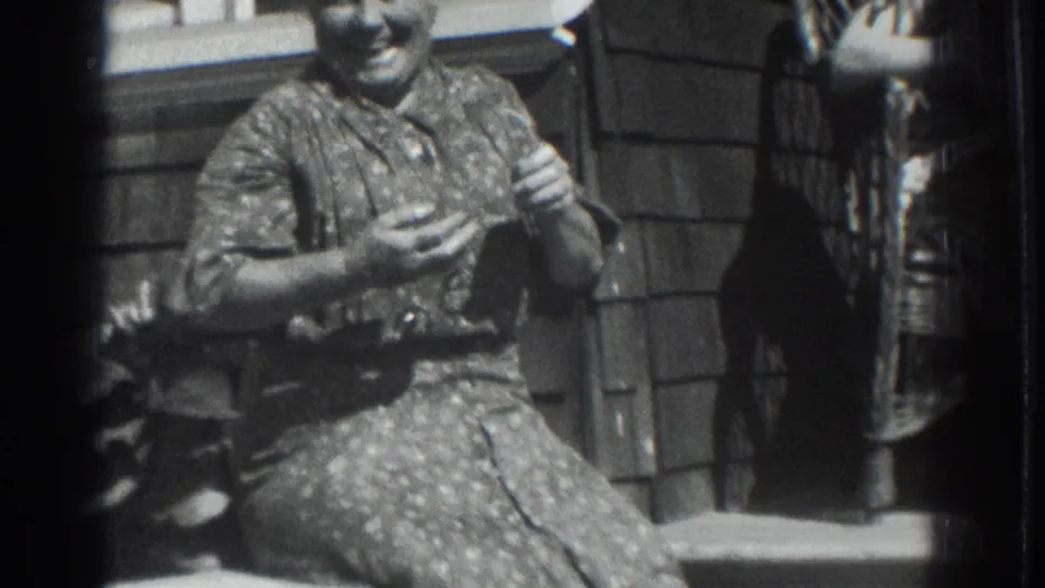 1937 Vacaville home-movie frame of an adult woman smiling on a porch while holding small knitting or handwork, with another woman cropped nearby.