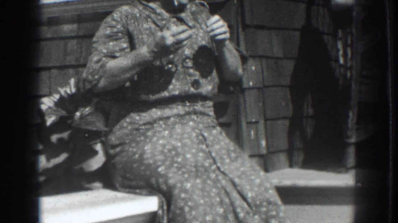 In 1937 Vacaville home-movie footage, a woman in a patterned dress sits on porch steps with hands near her face beside a shingled wall.