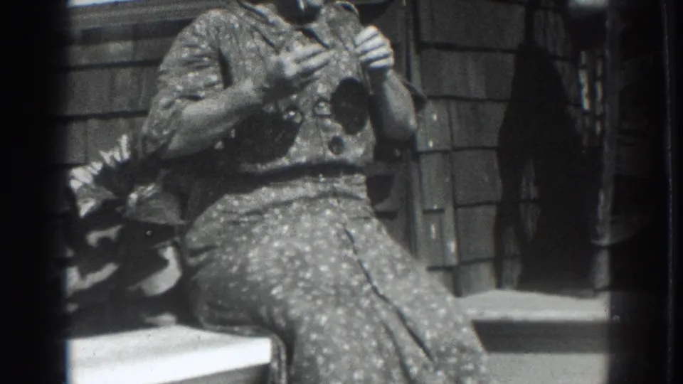 In 1937 Vacaville home-movie footage, a woman in a patterned dress sits on porch steps with hands near her face beside a shingled wall.