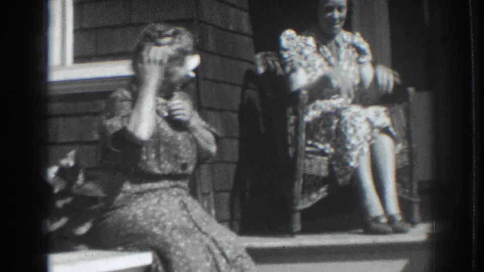 A woman seated on the porch steps raises a hand to her face as another woman in a patterned dress sits behind her in a wicker chair.