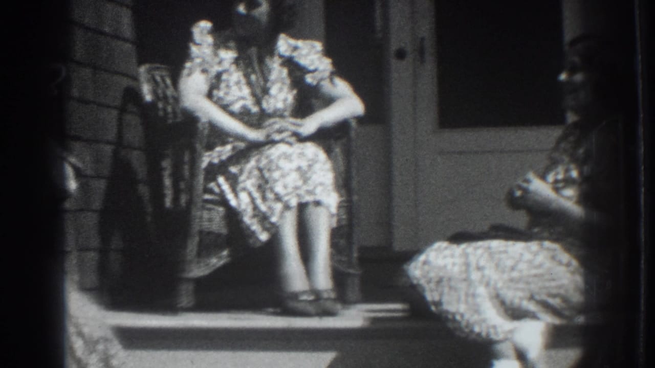 Two women sit on a porch, one in a wicker chair and one on the steps.