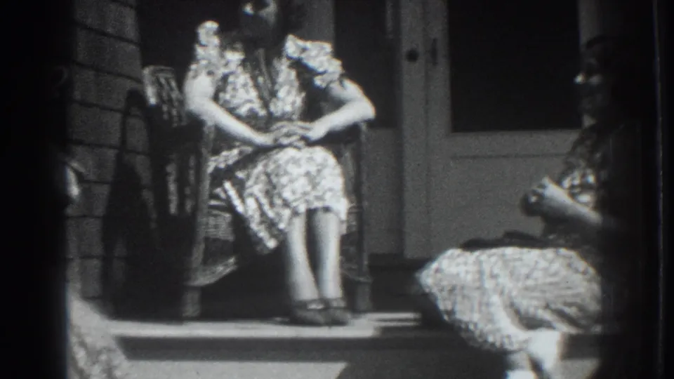 In 1937 Vacaville, two women in patterned dresses sit on a residential porch, one in a wicker chair and one partly turned on the steps.