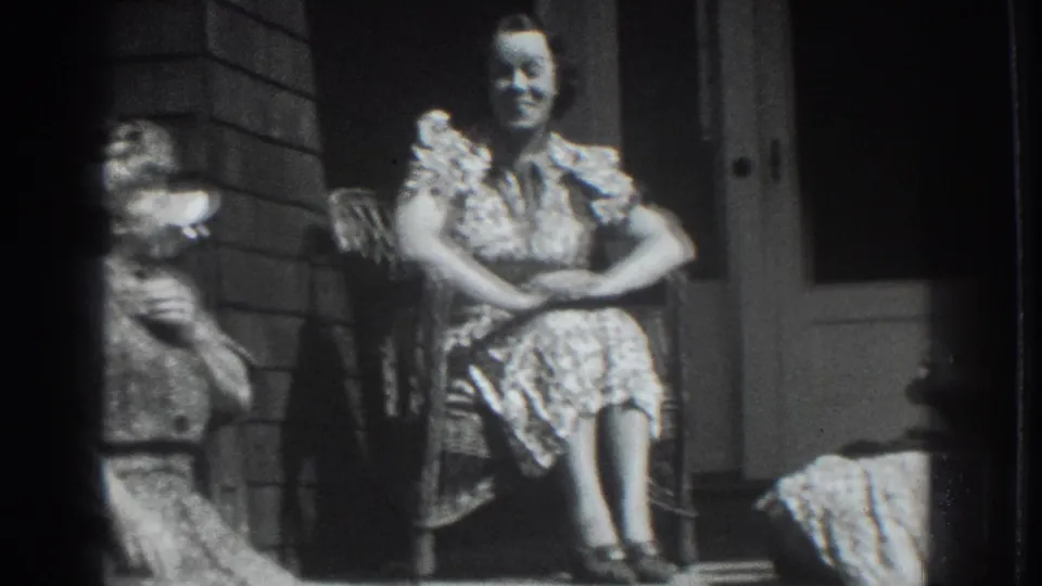 In 1937 Vacaville, a smiling woman sits in a wicker chair on a porch, with another seated figure partly visible at the lower edge.