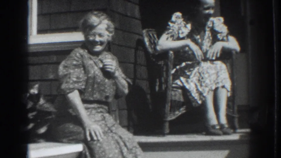In 1937 Vacaville, two women in patterned dresses sit on porch steps, one smiling toward the camera and the other seated higher near a chair.