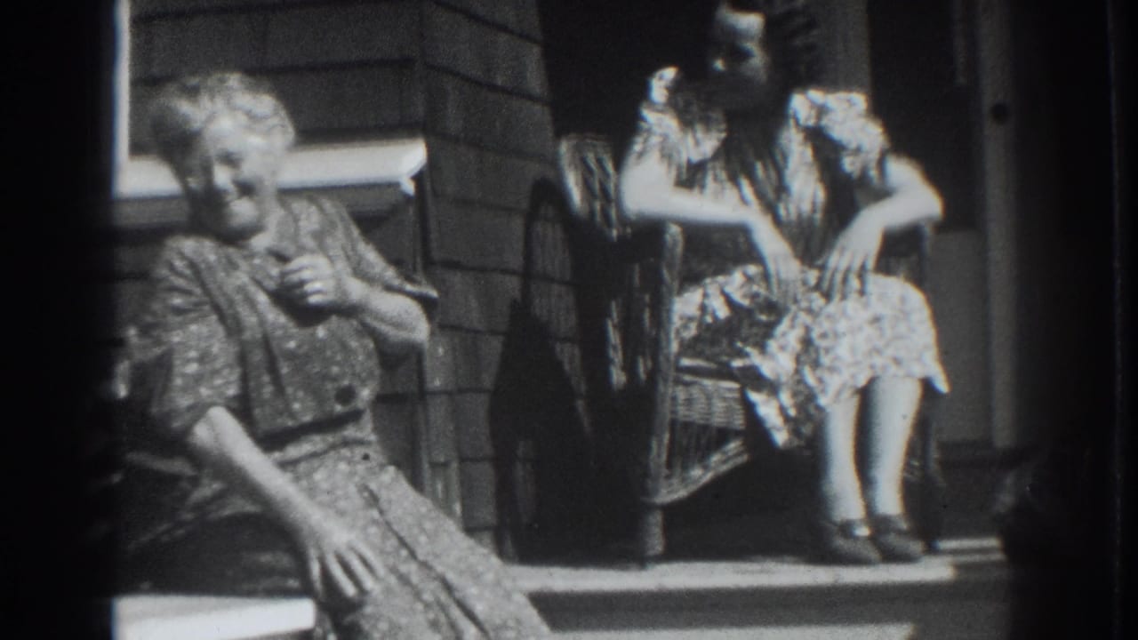 Two women in dresses sit on a porch beside a shingled wall.