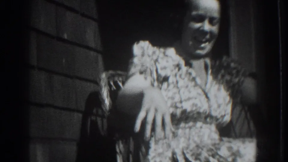 In this 1937 Vacaville home-movie frame, an adult woman smiles while seated in a wicker porch chair beside a shingled wall.