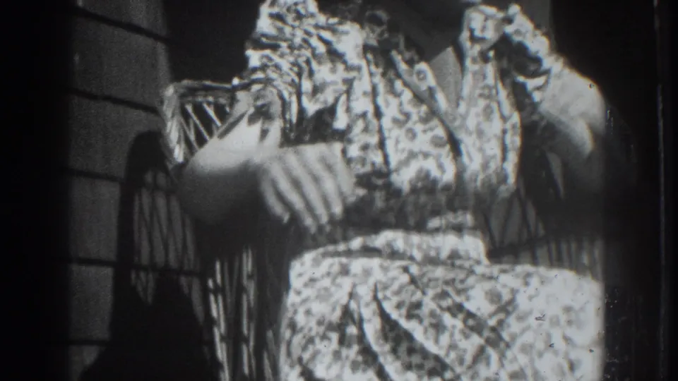 The seated woman remains partly framed in a wicker chair, with her patterned dress and porch wall visible in black-and-white 8mm footage.