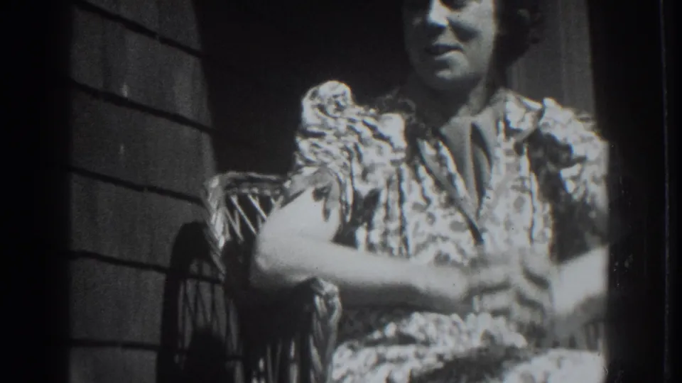 The woman sits in the wicker chair with her hands together, facing the camera in the close black-and-white home-movie view.