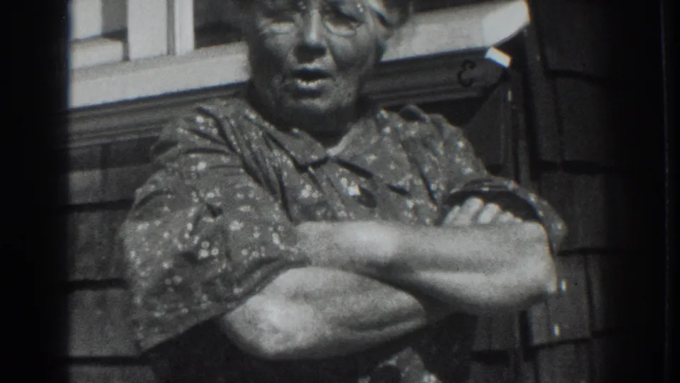 The 1937 black-and-white frame shows an older woman standing with arms crossed beside a house window in Vacaville.