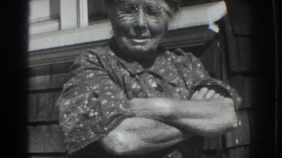 An older woman smiles with arms crossed in a black-and-white 8mm porch portrait from Vacaville, California, 1937.