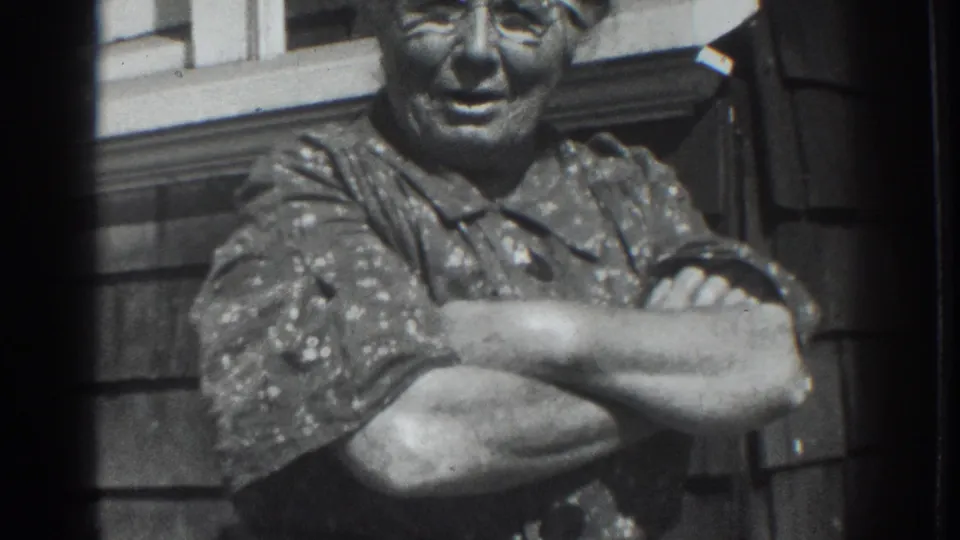 Older woman in 1937 Vacaville home-movie footage smiles with folded arms beside wood siding and a window.