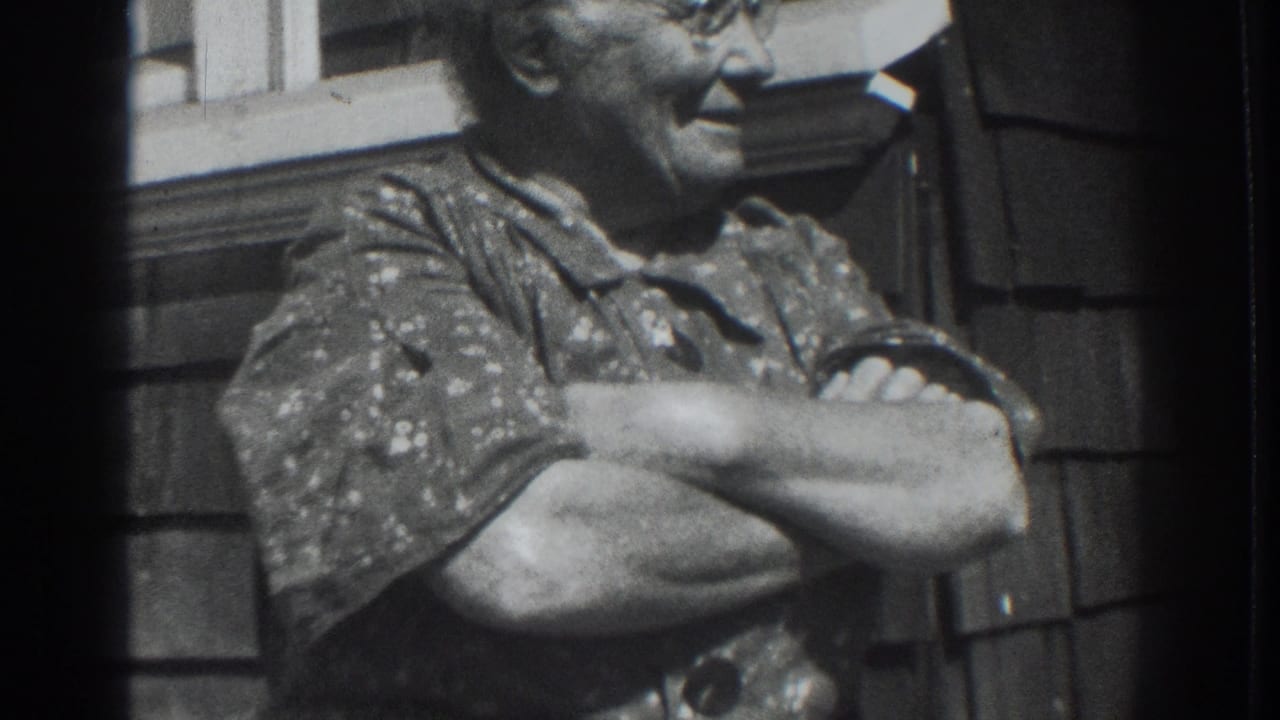 Older woman in glasses smiles with arms crossed outside a shingled house.