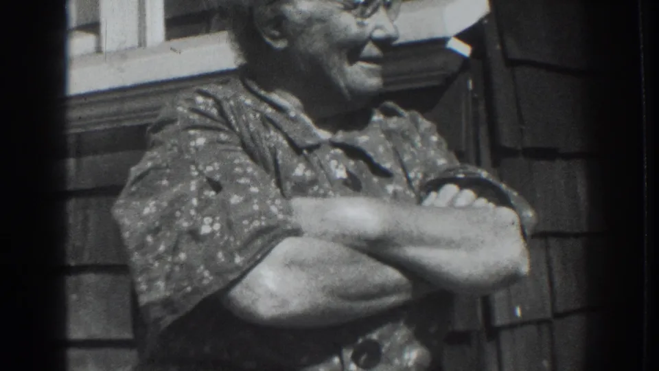 In Vacaville, California, 1937, an older woman in glasses and a patterned dress smiles with crossed arms beside shingle siding.