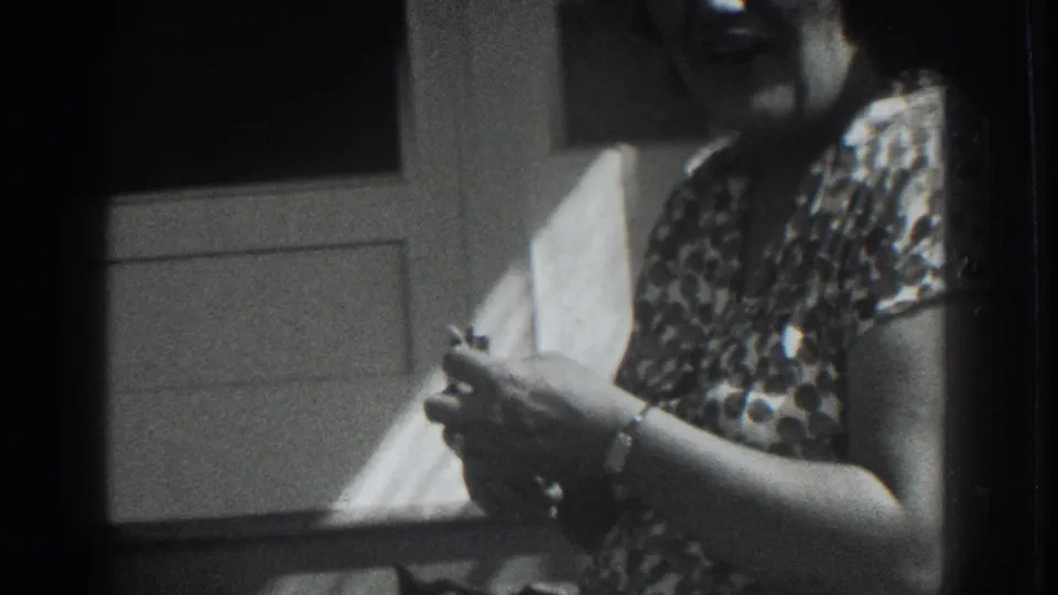 A woman in a patterned dress stands by a white door in the 1937 Vacaville home-movie scene, holding a small object in daylight.