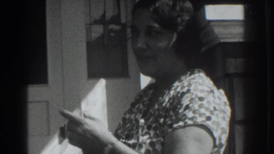 A woman in a patterned dress gestures with her hand beside the white door, appearing to address someone nearby in the 1937 Vacaville scene.
