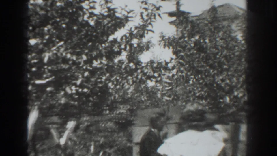 1937 Vacaville home-movie frame showing adults beneath fruit trees near boxes or a low fence in a backyard.