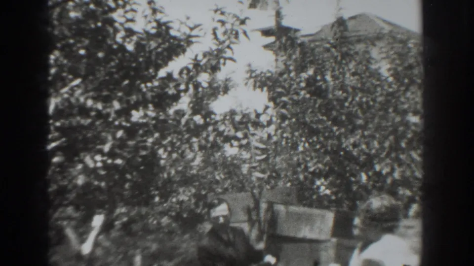 1937 Vacaville frame with dense tree branches foregrounding adults near backyard boxes.