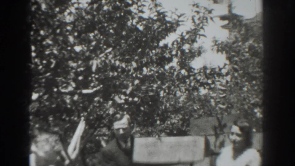 1937 Vacaville home-movie frame showing two adults in a leafy yard with a long board and soft black-and-white 8mm detail.