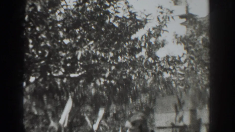 Black-and-white 8mm frame from Vacaville showing a leafy garden, an adult partly in view, and a board angled near the trees.