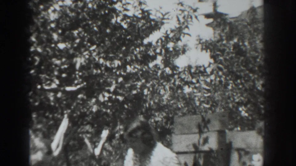 Black-and-white home-movie frame showing an adult in a Vacaville garden with a board, trees, and a low fence or wall behind.