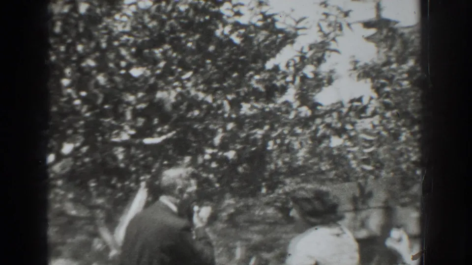 The man raises a hand or fruit near his face as the woman stands opposite him in the garden scene.