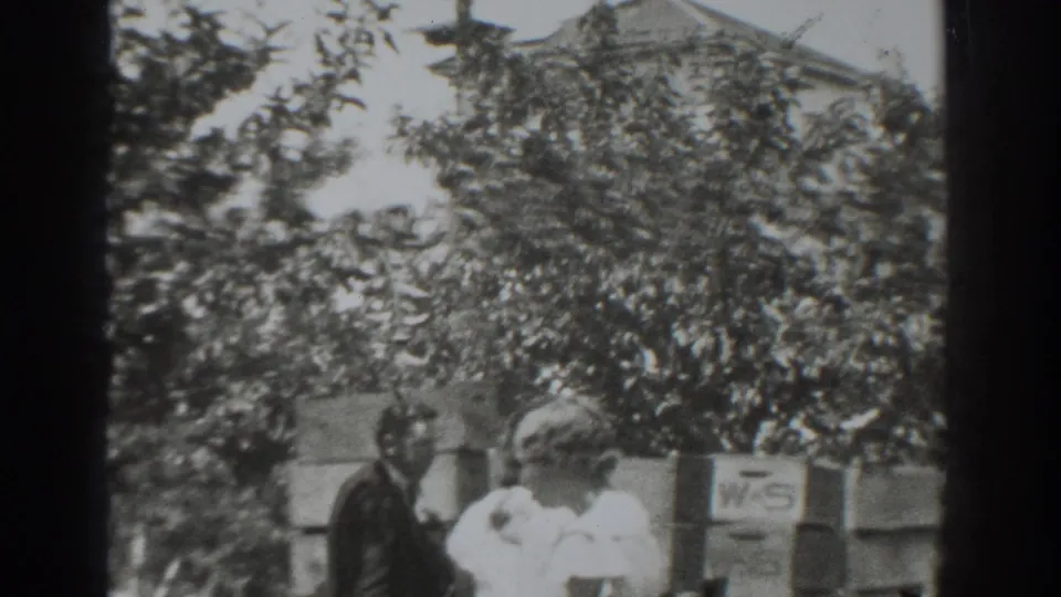 Adults in the Vacaville orchard stand near leafy trees, a low wall, a small sign, and a house roofline.
