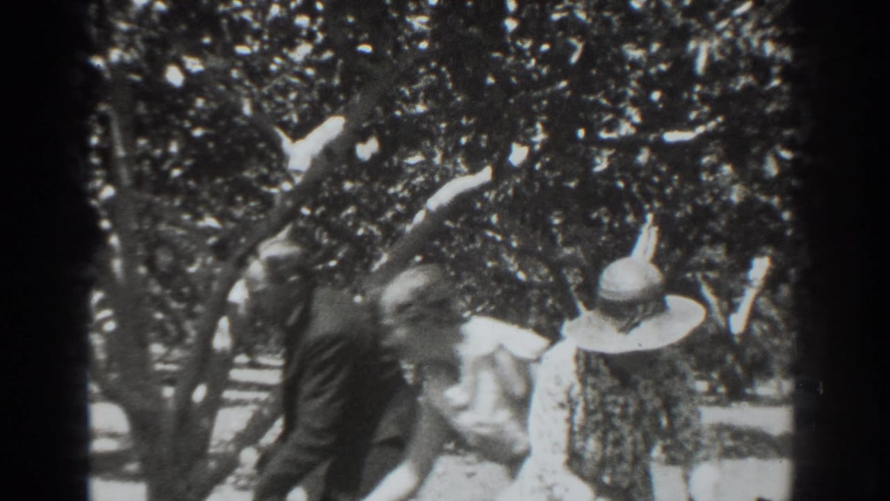 A man in a dark suit walks behind the woman in a wide-brimmed hat among orchard trees in the 1937 Vacaville footage.