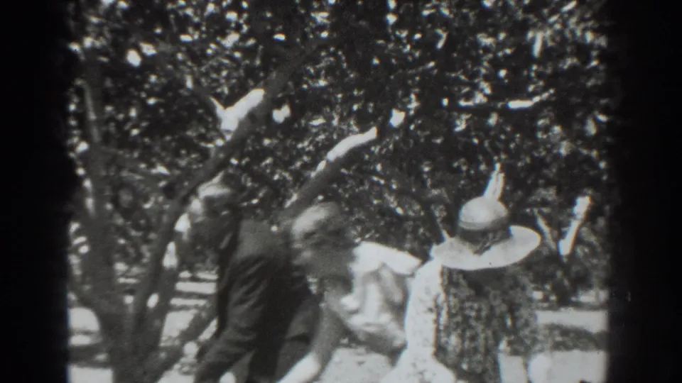 A man in a dark suit walks behind the woman in a wide-brimmed hat among orchard trees in the 1937 Vacaville footage.