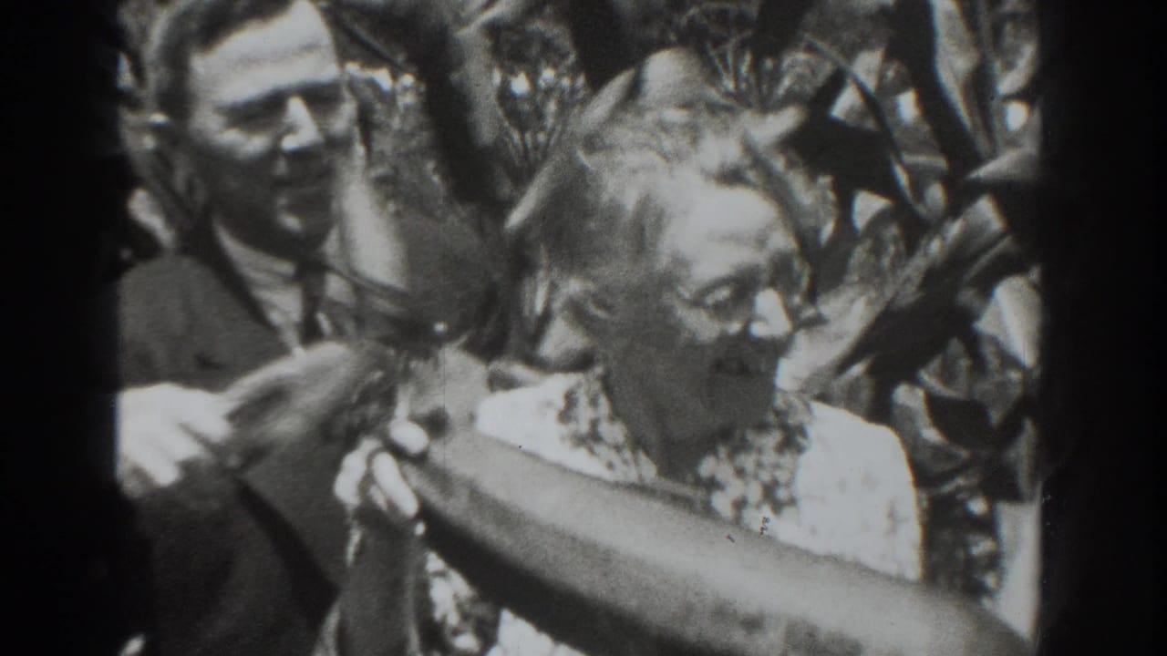 A man in a suit stands beside the older woman as she holds a long oversized vegetable in the Vacaville garden footage.