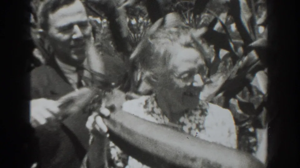 A man in a suit stands beside the older woman as she holds a long oversized vegetable in the Vacaville garden footage.