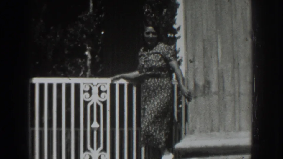 In the 1937 Vacaville home movie, a woman in a patterned dress poses beside a white porch rail and column.
