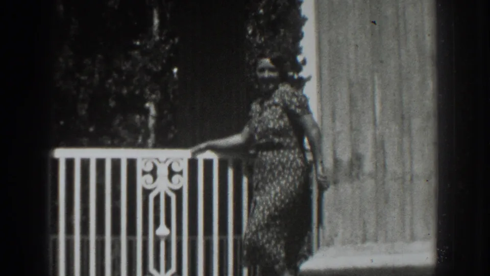 A woman stands beside the porch rail in the 1937 Vacaville footage, framed by a column, wall, and dark garden foliage.