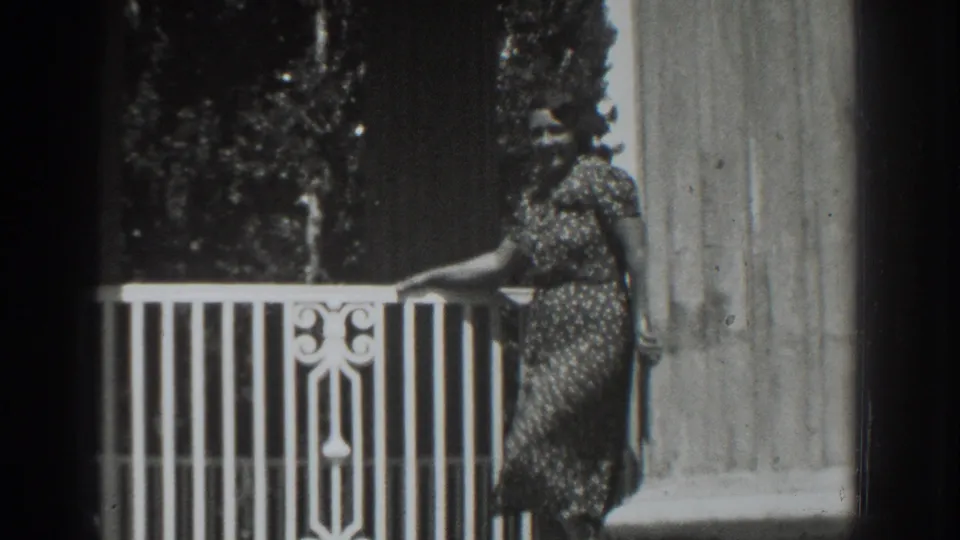 In 1937 Vacaville home-movie footage, a woman poses beside a white balcony rail, tall column, and dark foliage.