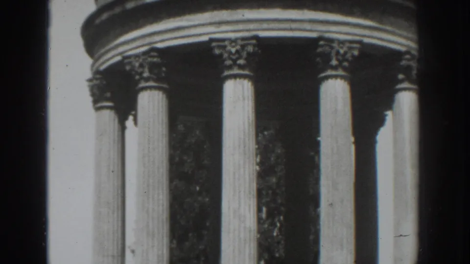 Black-and-white 8mm frame of the Vacaville columned structure, showing full column shafts, ornate capitals, and trees in the background.
