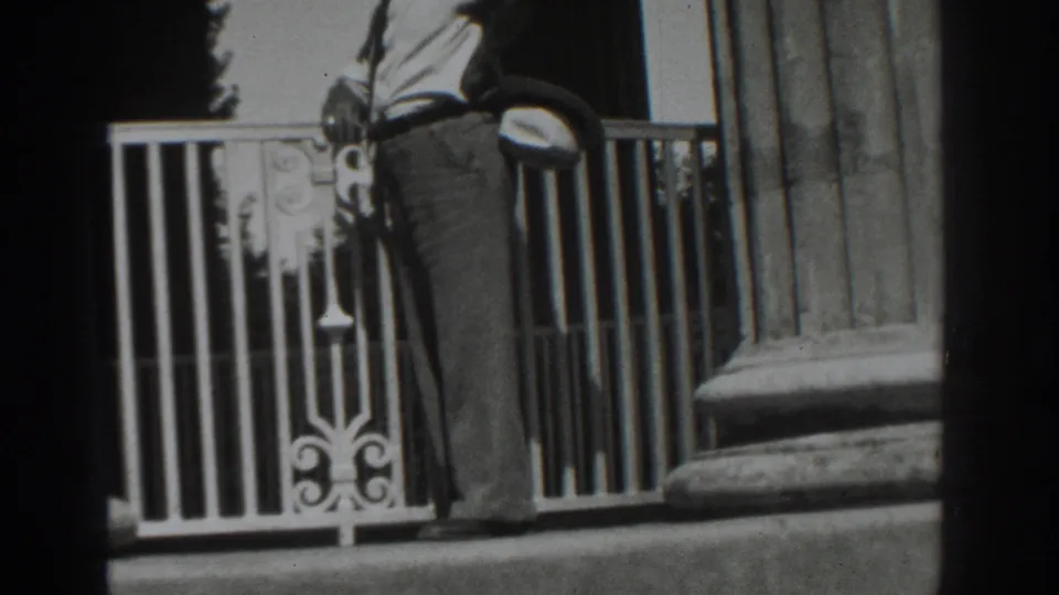 1937 Vacaville frame showing a man in period attire leaning near an ornate balcony railing by columns and steps.