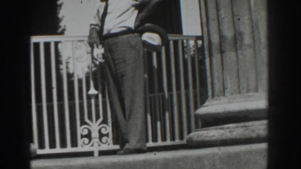 In 1937 Vacaville footage, a man in a light shirt and trousers stands by a white railing beside a large fluted column.