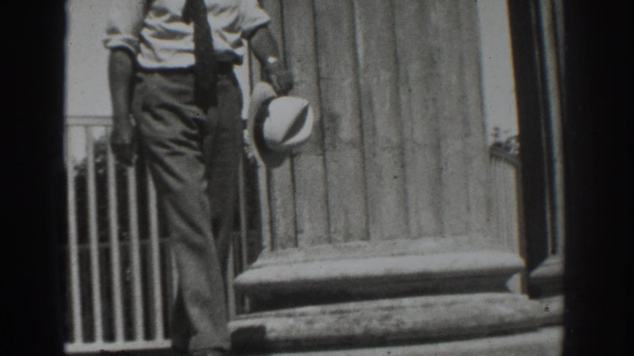 Man in shirt and tie steps down outside a columned building while holding a light-colored hat.