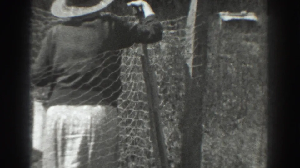 A woman in a wide-brimmed hat and long garment adjusts chicken wire near a wooden post in Vacaville, California, in 1937.