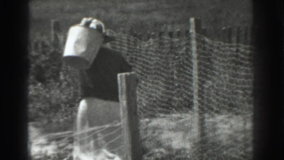A 1937 home-movie frame shows the woman passing a fence post while carrying the metal bucket on sloped ground.