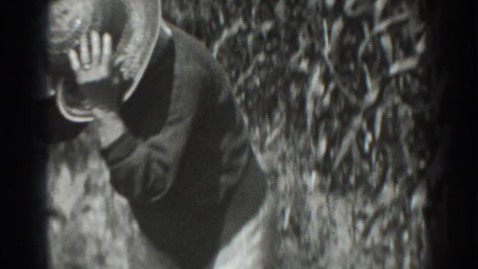 The 8mm frame shows the worker close to the camera, partly turned, with a hand near the brim of a field hat.