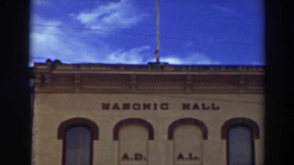 1937 color 8mm frame showing the Virginia City Masonic Hall facade below blue sky, clouds, and roofline detail.